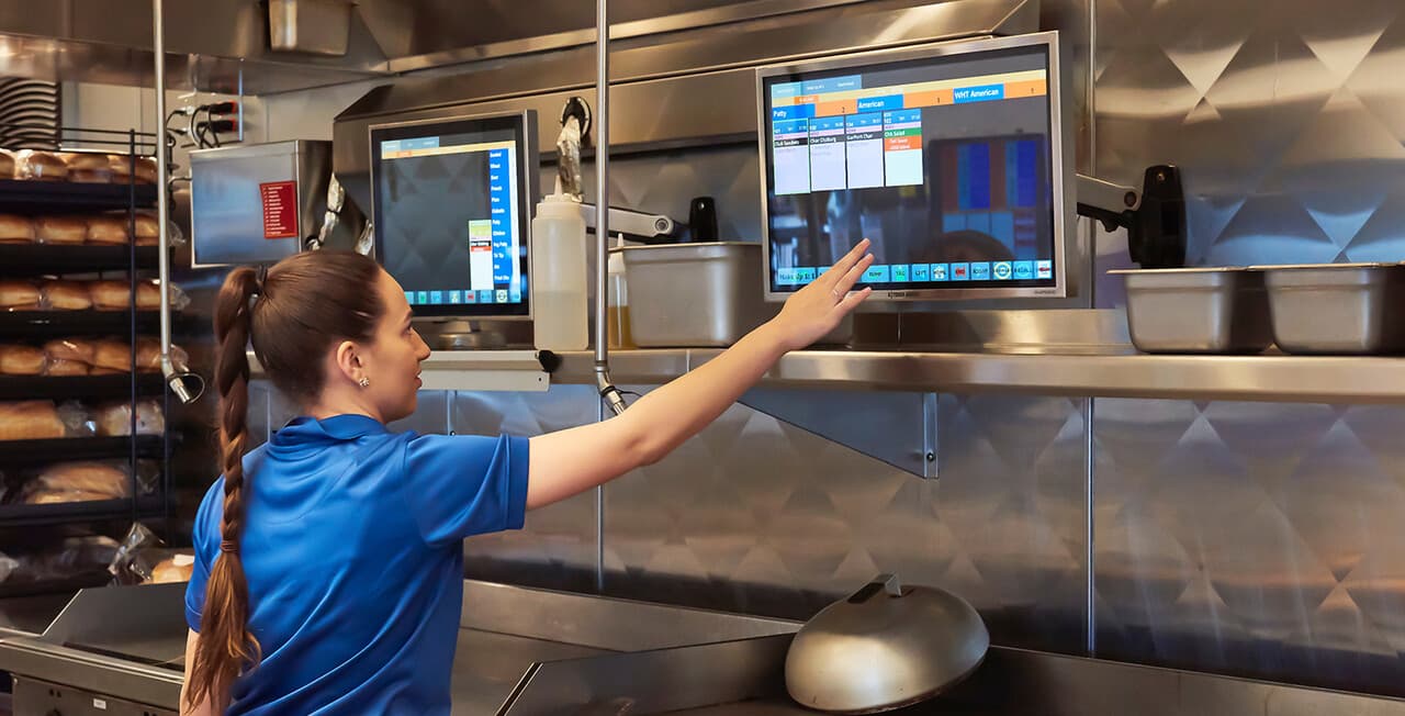 Restaurant staff member using a wall-mounted touchscreen kitchen display system to track and route orders in a high-volume commercial kitchen.